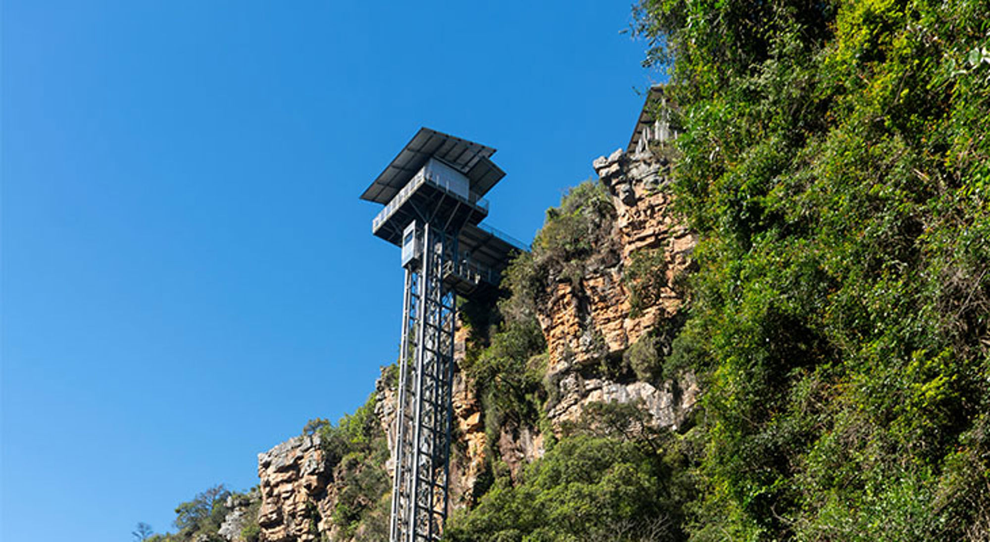 Uitzichtplatform en lift bij de Tsitsikamma-kloof in Zuid-Afrika, met rotswand en groen tegen een blauwe lucht.