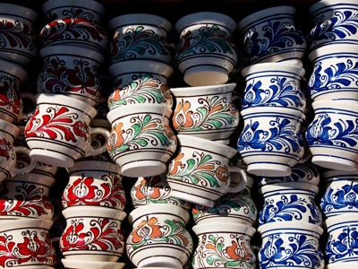 Handbeschilderde Roemeense keramiekbekers met bloemmotieven in rood, groen en blauw, gestapeld op een markt.