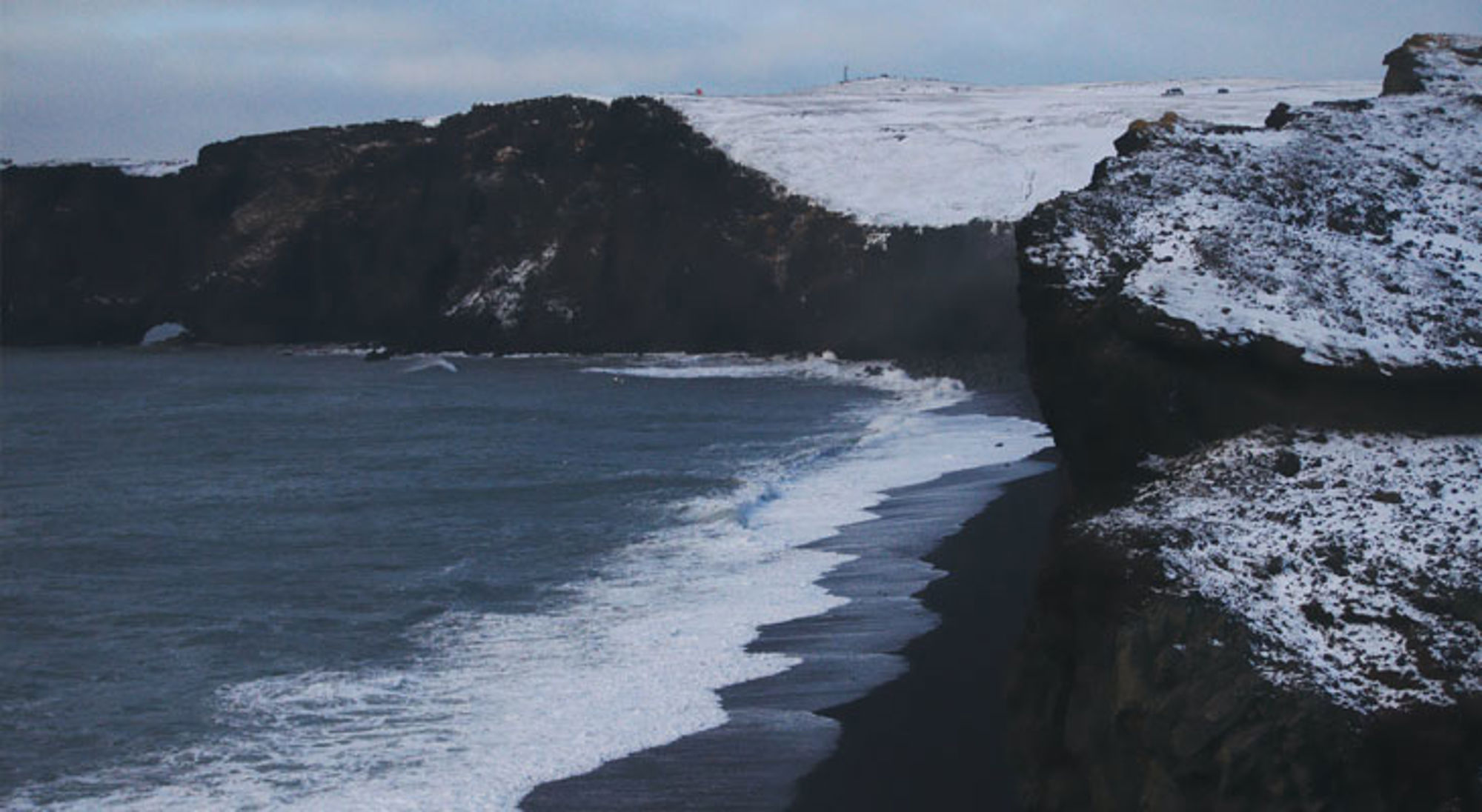 Zwarte zandkust met besneeuwde kliffen en golven aan de zuidkust van IJsland.
