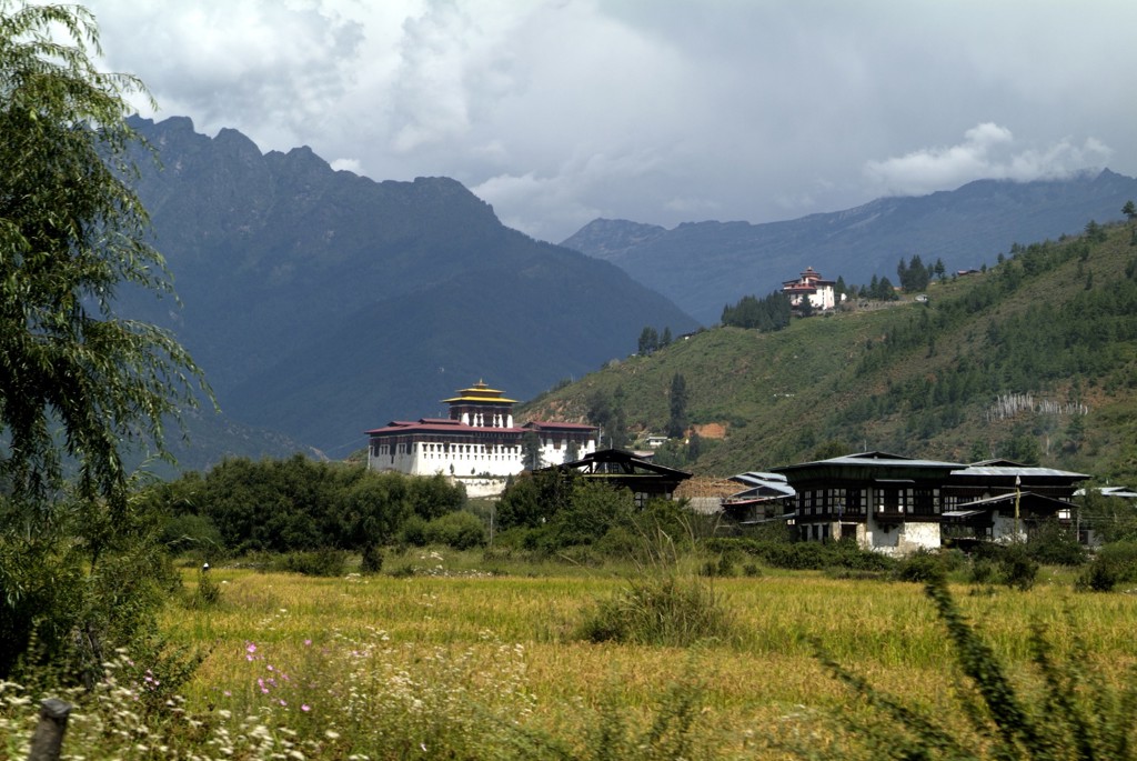 Bhutaans klooster en traditionele huizen in een groene vallei, met Himalaya-bergen op de achtergrond.