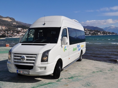 Witte minibus voor vervoer tijdens Balkanreizen, geparkeerd aan de kust met bergen op de achtergrond.
