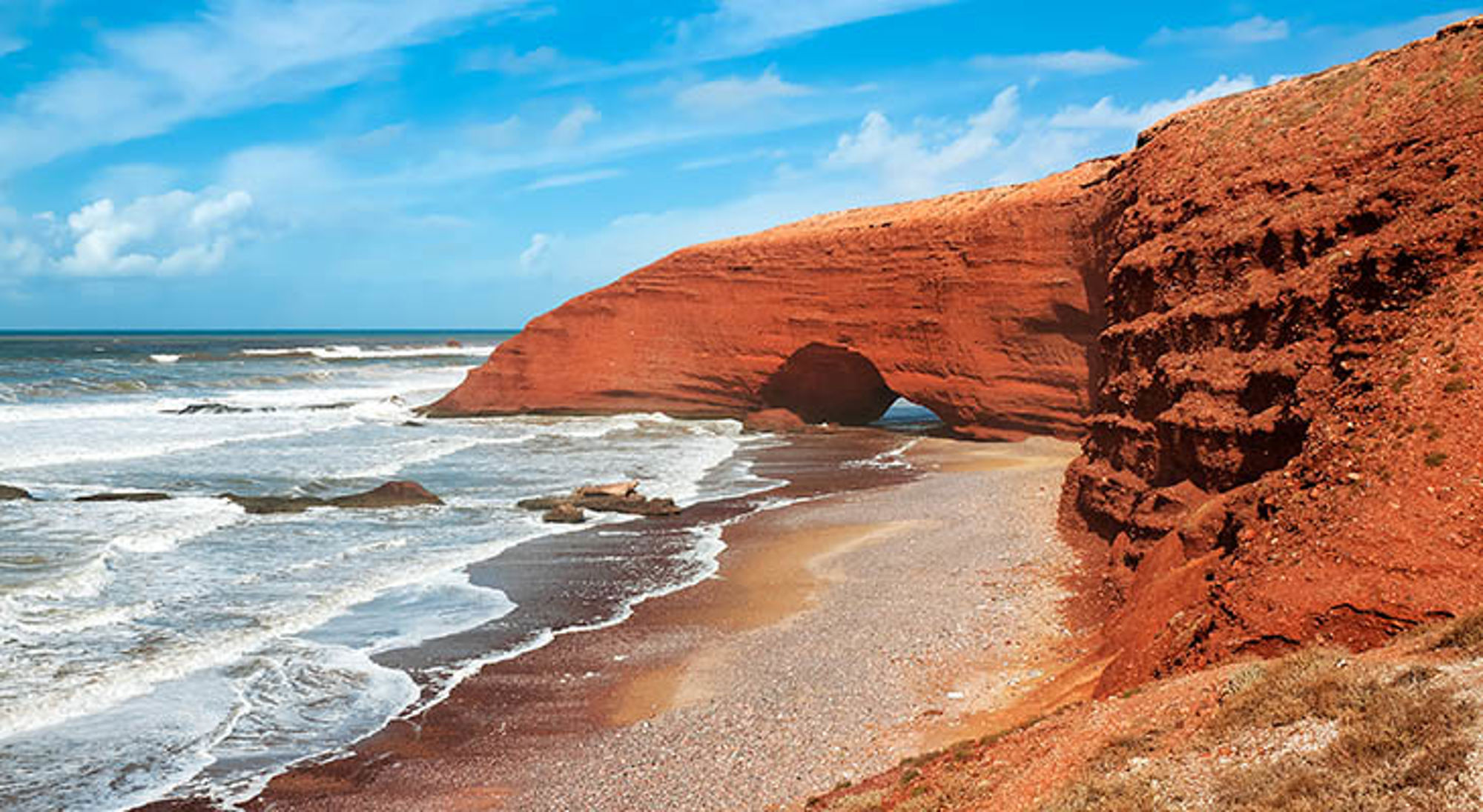 Rode rotsboog aan de Atlantische kust van Marokko, met golven en zandstrand onder een blauwe lucht.
