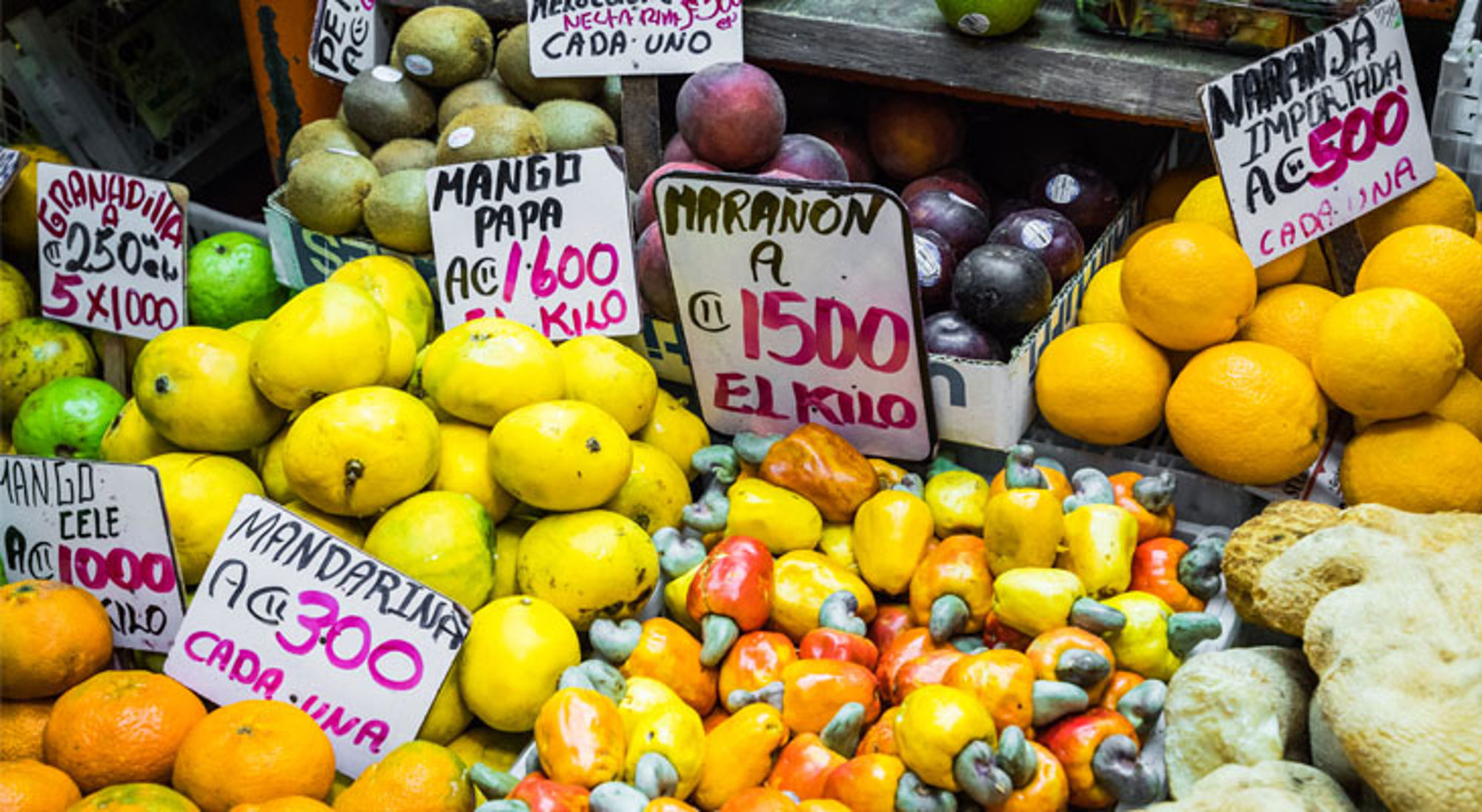 Kleurrijke fruitkraam op een markt in Costa Rica met mango’s, mandarijnen, cashewnoten en prijsbordjes.