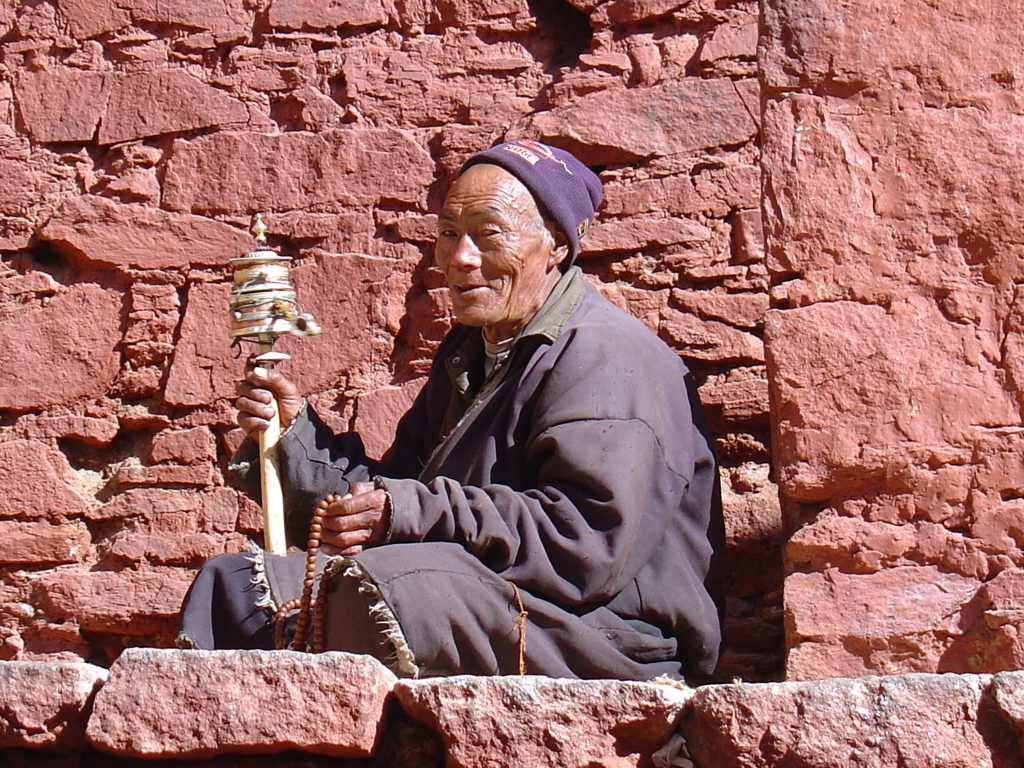 Tibetaanse man met gebedsmolen en mala, zittend tegen een roodstenen muur tijdens groepsrondreis Tibet en Nepal.