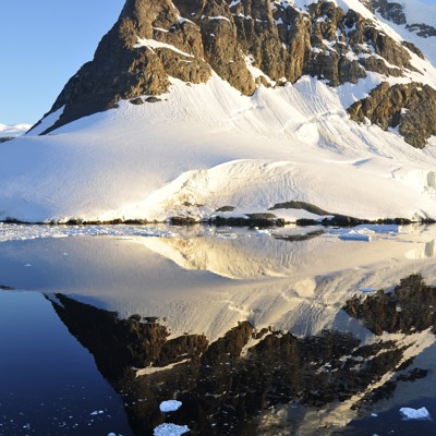 Besneeuwde bergen en gletsjerkust in Antarctica, weerspiegeld in kalm water met drijvend ijs.
