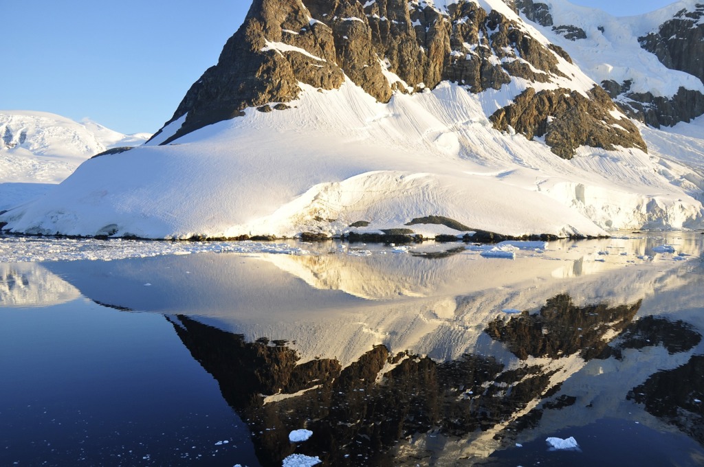 Besneeuwde bergen en gletsjerkust in Antarctica, weerspiegeld in kalm water met drijvend ijs.