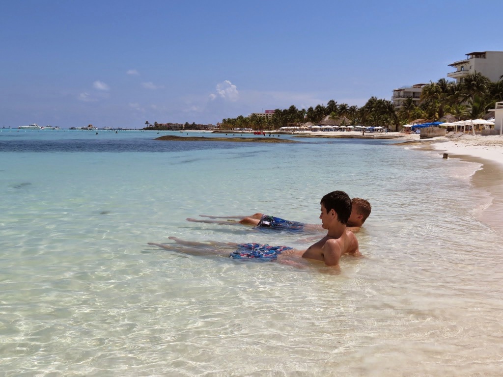 Kinderen ontspannen in ondiep zeewater aan een wit zandstrand in Mexico tijdens een familiereis.
