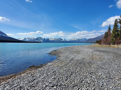 Kiezelstrand aan een helderblauw meer met besneeuwde bergen op de achtergrond in Alaska.