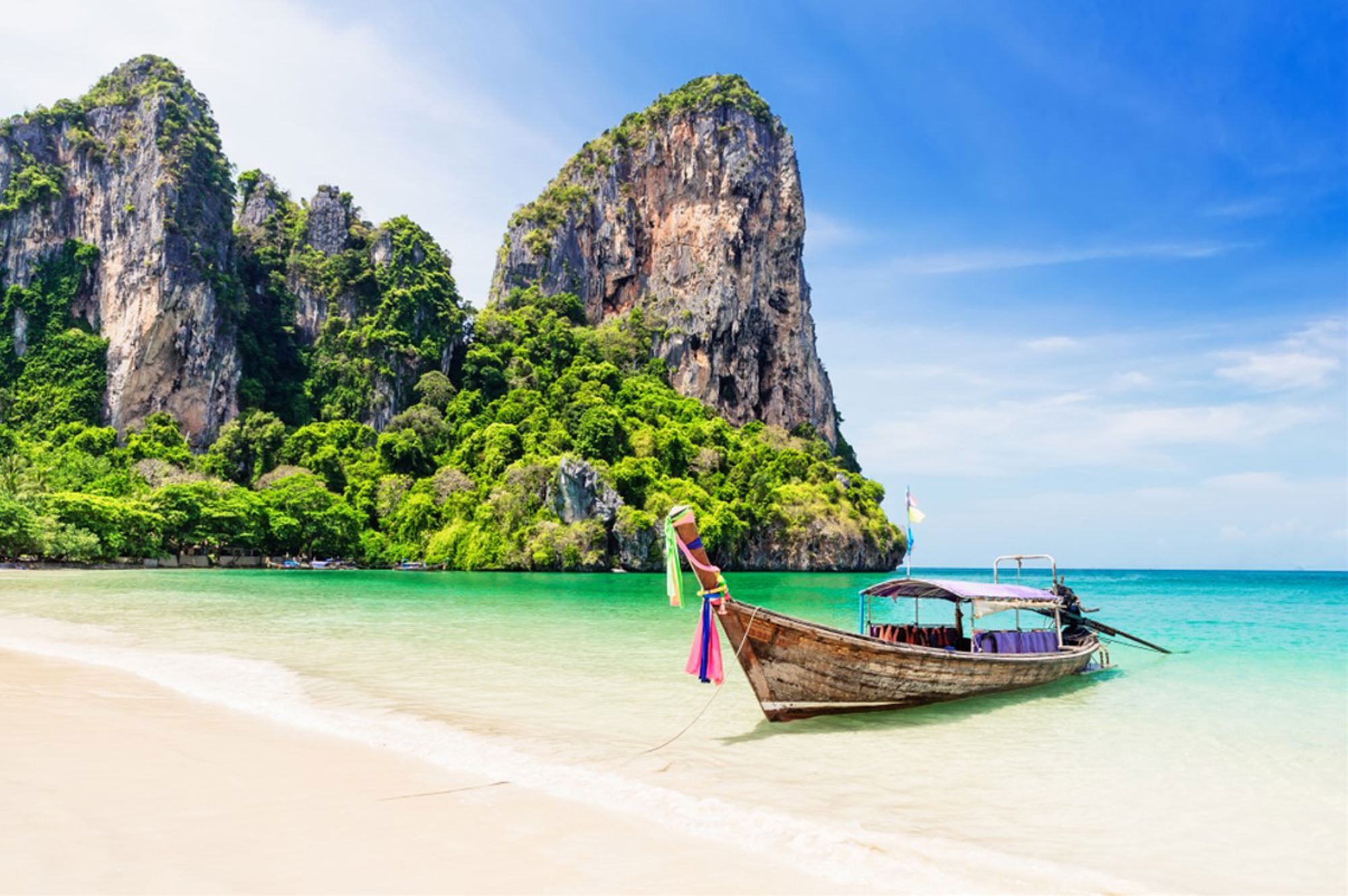 Longtailboot bij een wit zandstrand met kalkstenen kliffen en turquoise zee in Thailand.