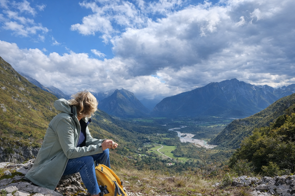 Wandelaar met rugzak kijkt uit over een bergvallei in Slovenië, met rivier en bergen onder een dramatische lucht.