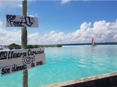 Bordje bij Bacalar-lagune met turquoise water en zeilboot in de verte, tijdens de familiereisroute Mexico.