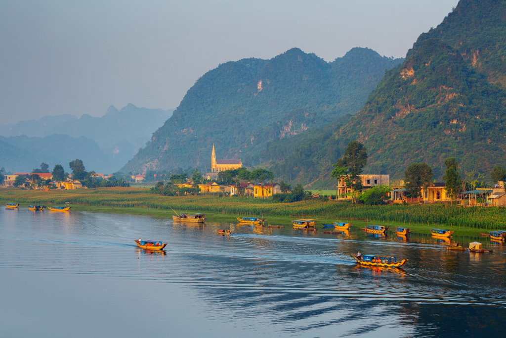 Rivierlandschap in Vietnam met houten boten, rijstvelden en groene karstbergen bij een dorp.