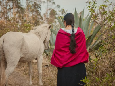 Vrouw in traditionele kleding met rode omslagdoek naast een wit paard in het landschap van Ecuador.