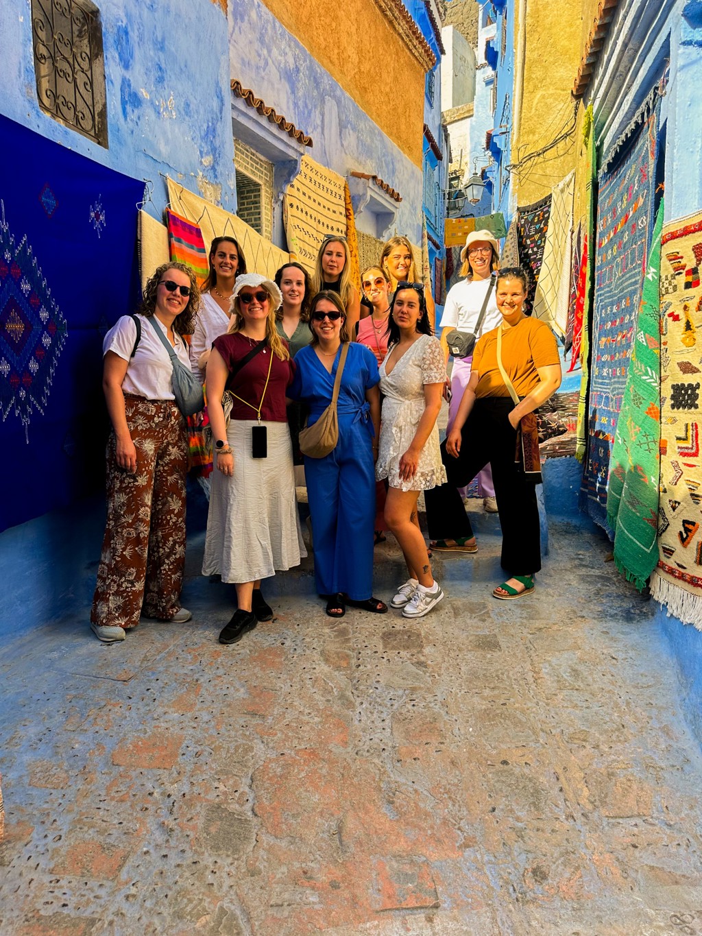 Groep reizigers tijdens 22-35ers reis Marokko in een smalle, blauw geschilderde straat met kleurrijke tapijten.