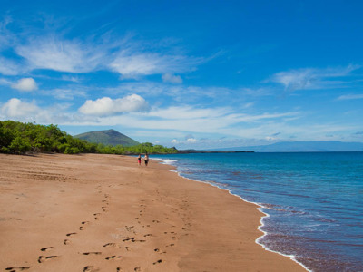 Wandelaars op een verlaten zandstrand op de Galapagoseilanden, met blauwe zee en vulkanische heuvels op de achtergrond.