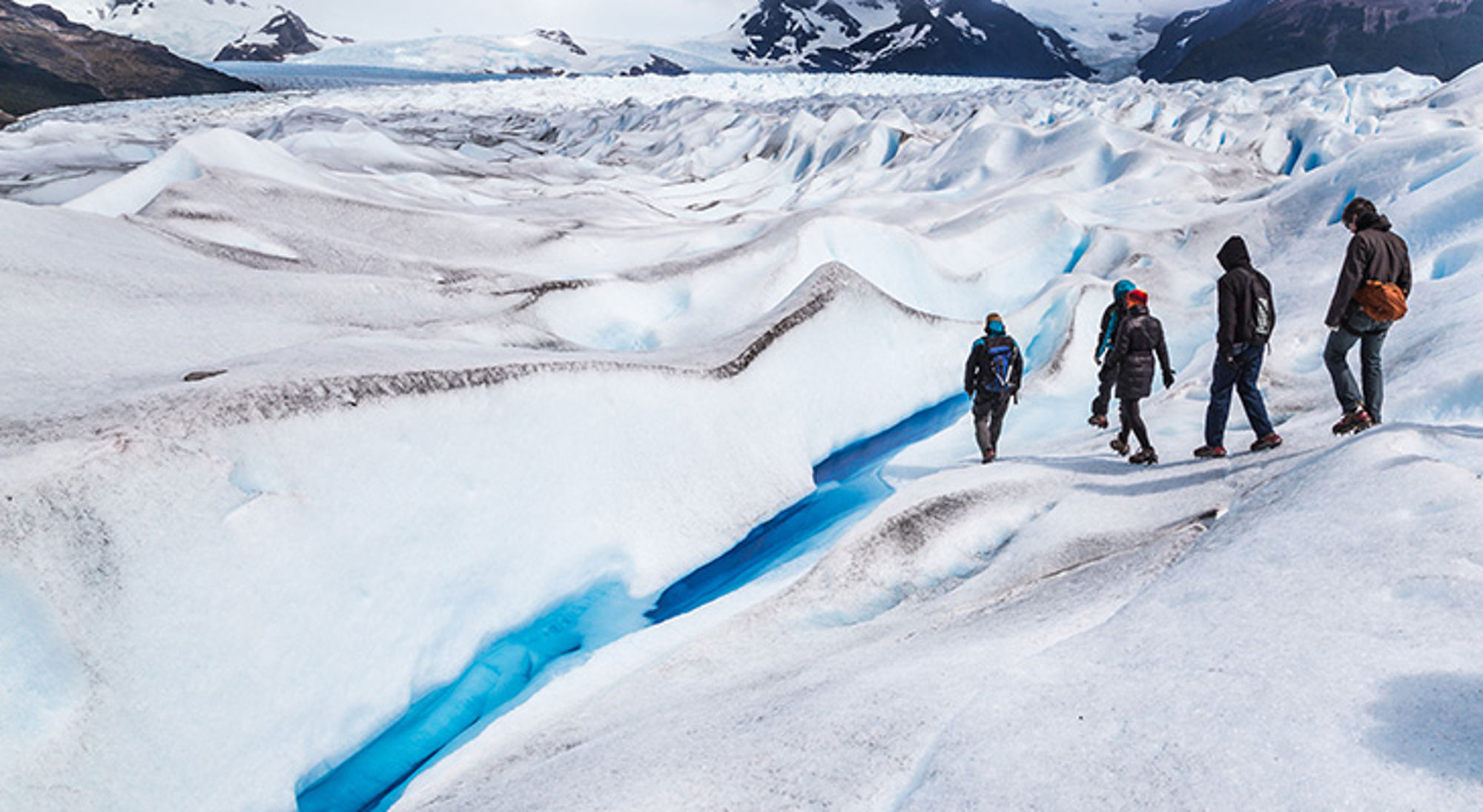 Wandelaars op een gletsjer in Patagonië, met diepe blauwe ijsspleten en besneeuwde bergen op de achtergrond.