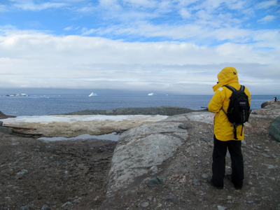 Reiziger in gele jas kijkt uit over de Antarctische kust met zee en ijsbergen tijdens Route Antarctica.