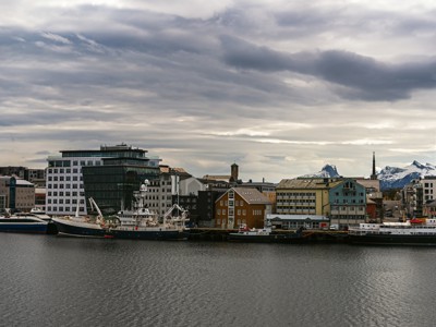 Havenstad in Noorwegen met aangemeerde schepen, moderne gebouwen en besneeuwde bergen op de achtergrond.