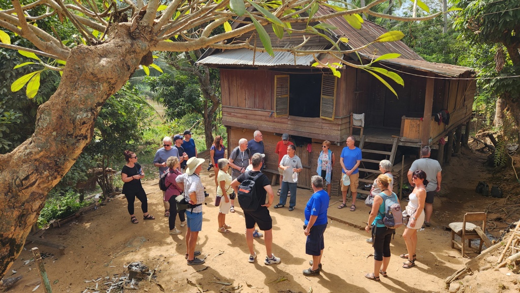 Groepsrondreis Laos & Cambodja: reizigers bezoeken een dorp en luisteren naar uitleg bij een houten huis op palen.