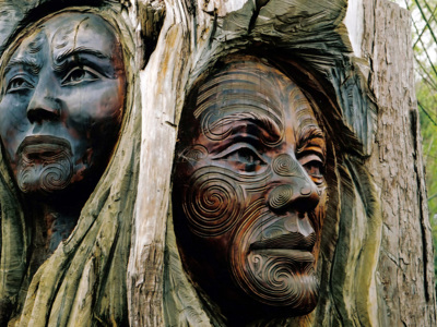 Houten Maori-beeldhouwwerk met twee gezichten en traditionele tatoeagepatronen langs de Route Nieuw-Zeeland.