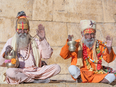 Twee sadhu’s met beschilderde gezichten zitten voor een stenen muur en zegenen met opgeheven hand.