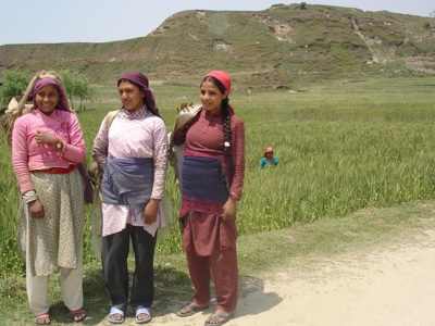 Drie vrouwen in traditionele kleding bij een rijstveld in Nepal, met groene heuvels op de achtergrond.