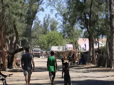 Straatbeeld in Madagascar met wandelaars, lokale kraampjes en een vrachtwagen onder hoge bomen.