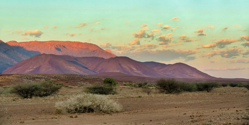 Berglandschap in Namibië bij zonsondergang, met rode heuvels en savannevegetatie onder een wolkenlucht.