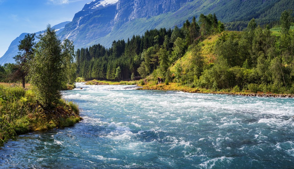 Berglandschap in Noorwegen met snelstromende rivier, groene bossen en besneeuwde toppen.