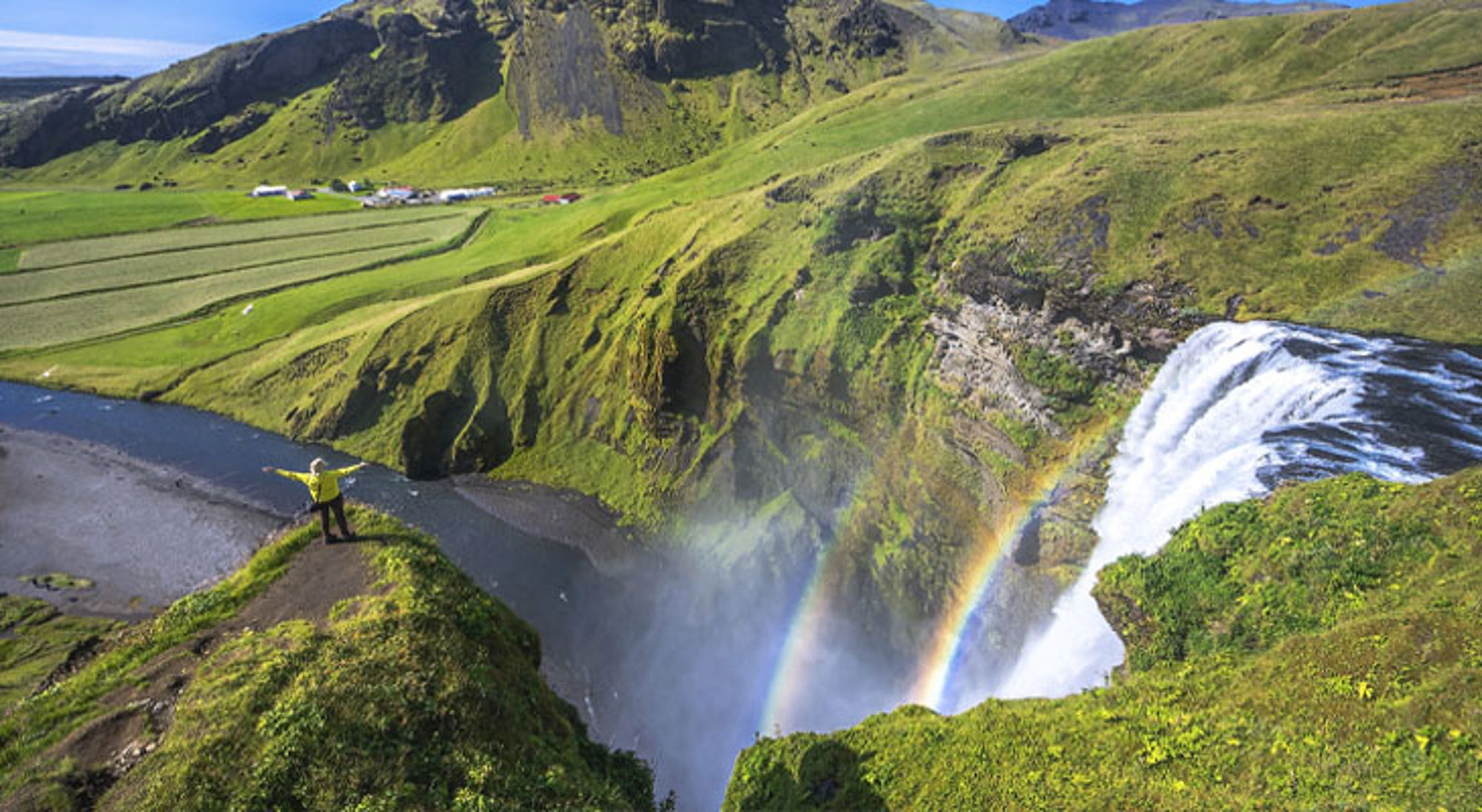 Waterval Skógafoss in IJsland met regenboog in de nevel, omringd door groene heuvels.