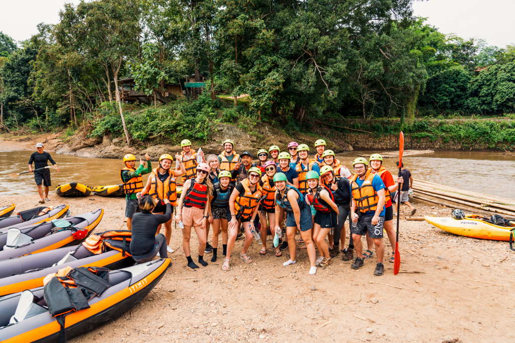 Groep 22-35ers klaar voor rafting op een rivier in Thailand, met helmen en reddingsvesten op de oever.