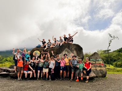 Groepsreizigers poseren bij het Arenal 1968-bord op de route Costa Rica, met groene heuvels en wolkenlucht.