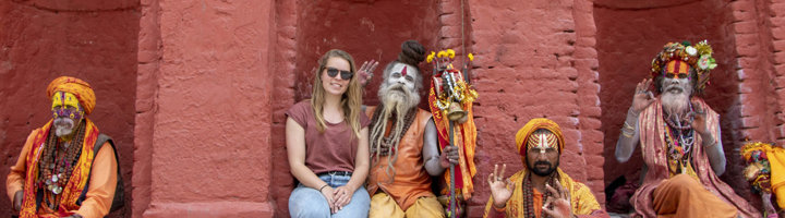 Vrouw op groepsreis poseert met kleurrijk geklede sadhu’s tegen een rode muur.