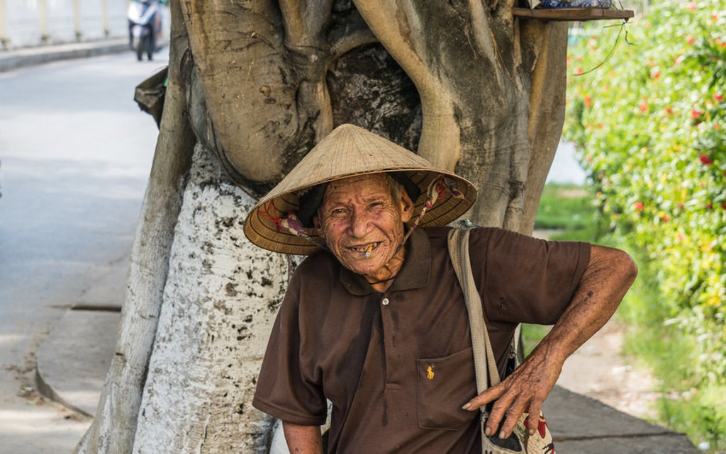 Vietnamees met traditionele kegelvormige hoed leunt tegen een boom langs de weg.
