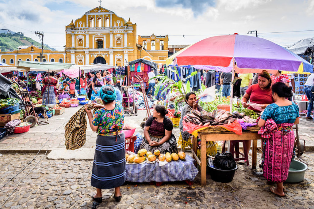 Drukke markt in Guatemala met kraampjes onder parasols en een gele kerk op de achtergrond.
