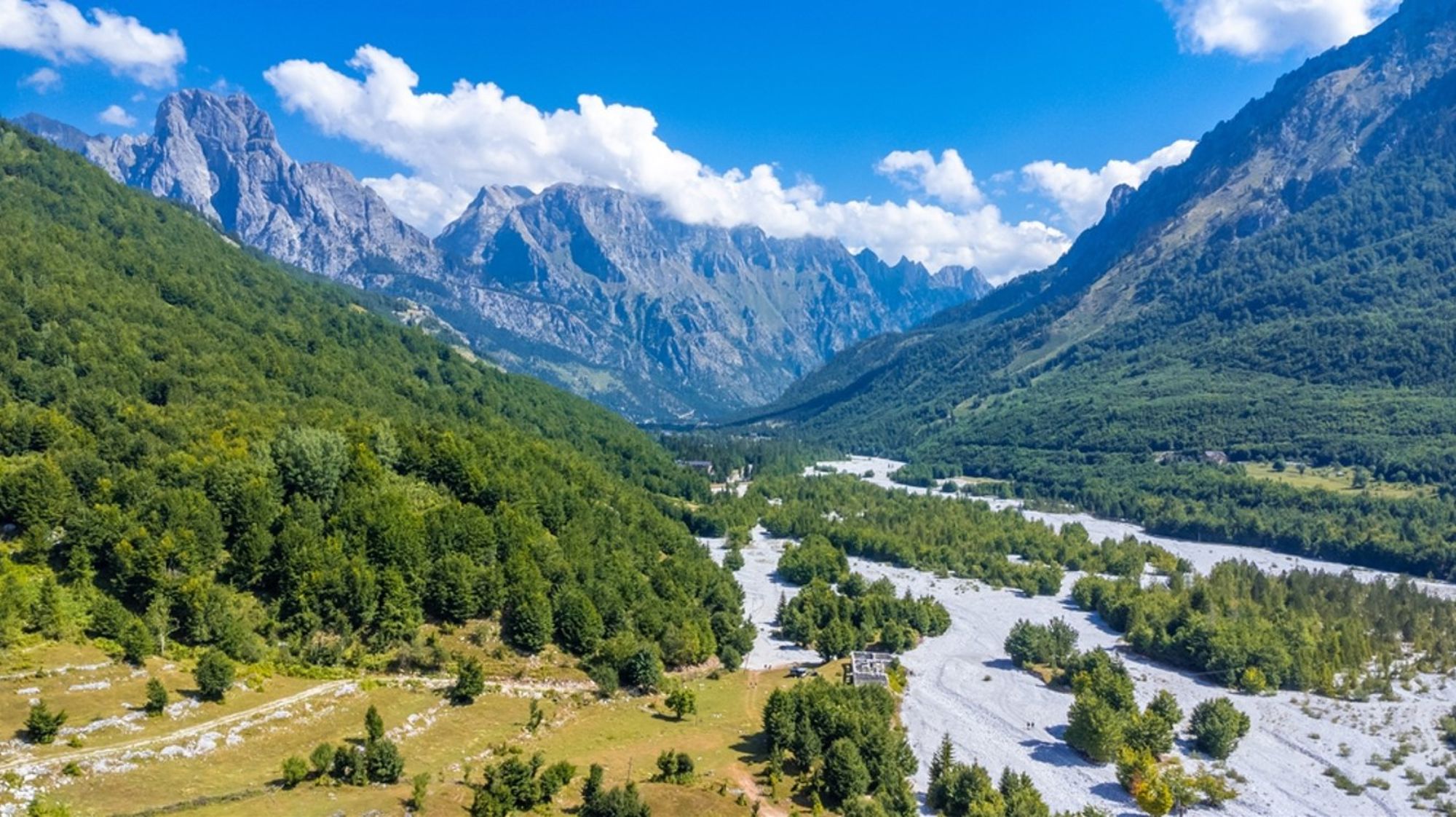 Valbonë-vallei in de Albanese Alpen met rivierbedding, groene bossen en ruige bergtoppen onder blauwe lucht.