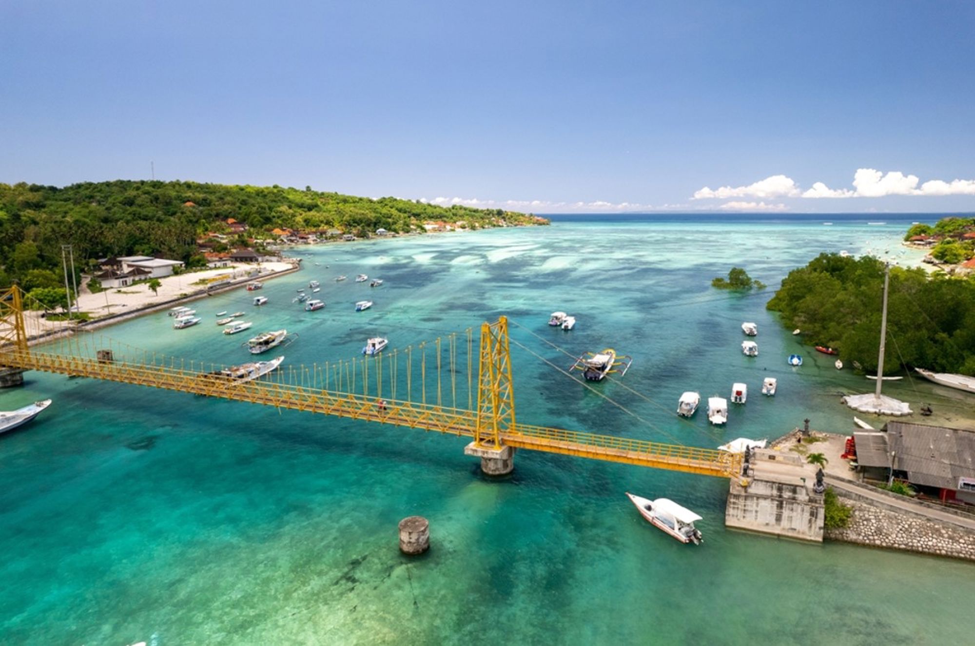 Gele hangbrug over een turquoise lagune met boten bij een Indonesisch eiland.