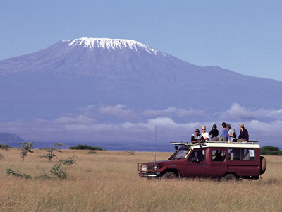 Safari-jeep met reizigers op het dak in de savanne, met de besneeuwde Kilimanjaro op de achtergrond.