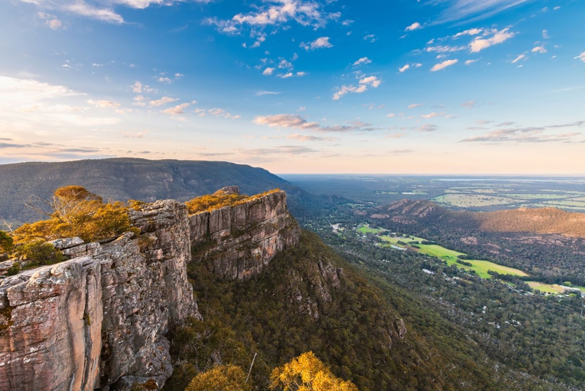Uitzicht vanaf een rotsklif over een groene vallei en berglandschap in Australië bij zonsondergang.