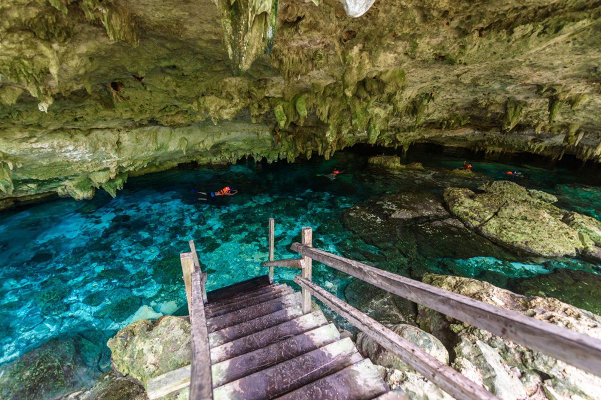 Trap naar een kristalheldere cenote in een grot in Mexico, met snorkelaars in turquoise water.