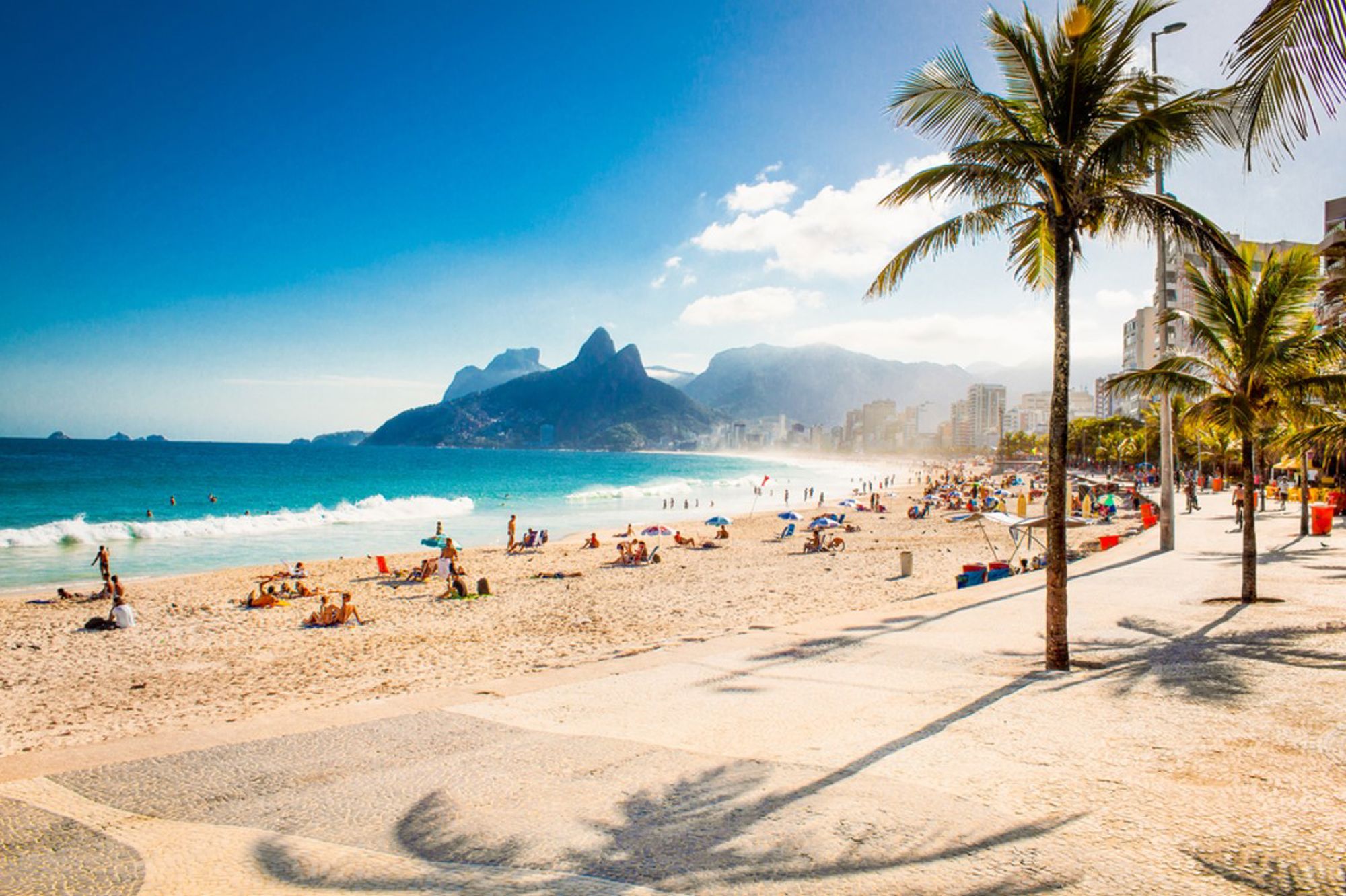 Zonnig strand van Ipanema in Rio de Janeiro met palmbomen, boulevard en bergen op de achtergrond.