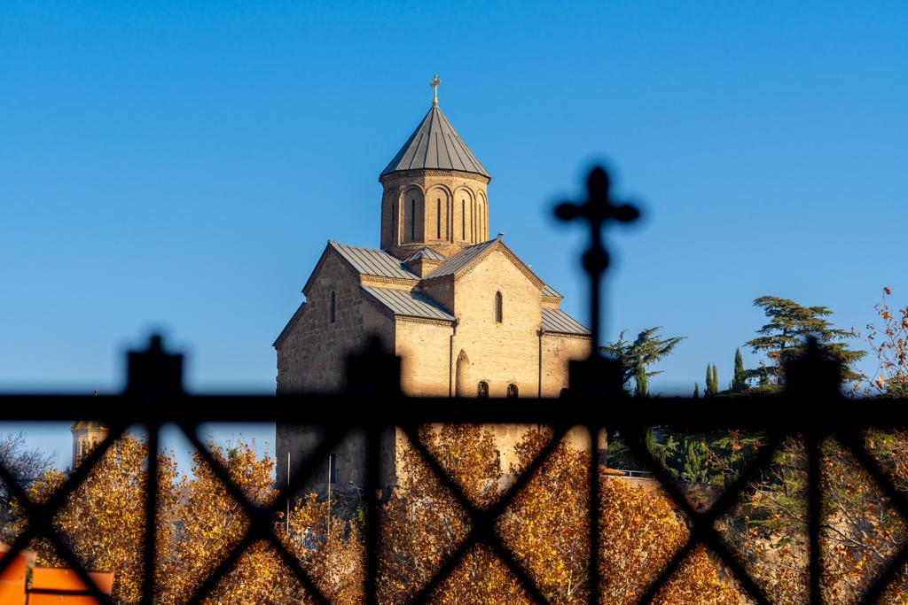 Georgisch orthodoxe kerk met koepel tegen een heldere blauwe lucht, gezien door een smeedijzeren hek.