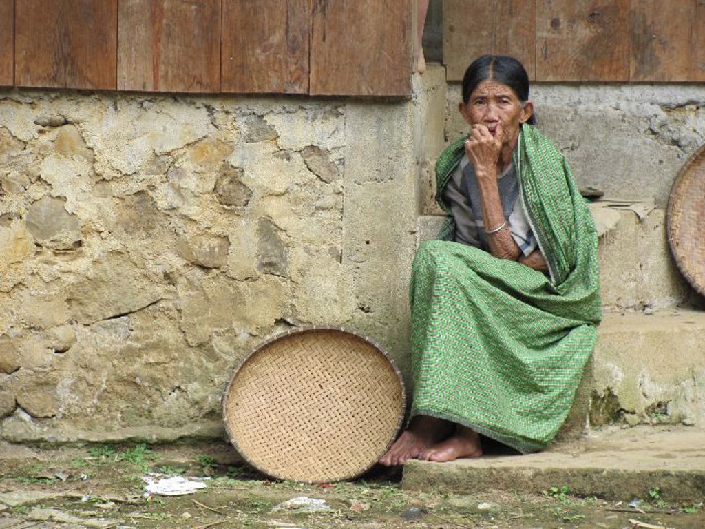 Vrouw in traditionele kleding zit bij een houten huis op Sulawesi, met een rieten mand naast haar.