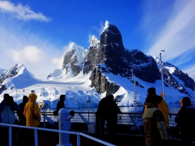 Reizigers op het dek tijdens de route Antarctica, vliegen en varen, met besneeuwde bergen op de achtergrond.