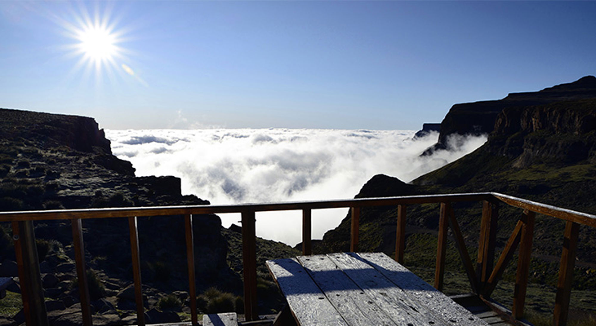 Uitzicht vanaf een houten terras over een wolkenzee tussen bergen in Zuid-Afrika, met de zon hoog aan de hemel.
