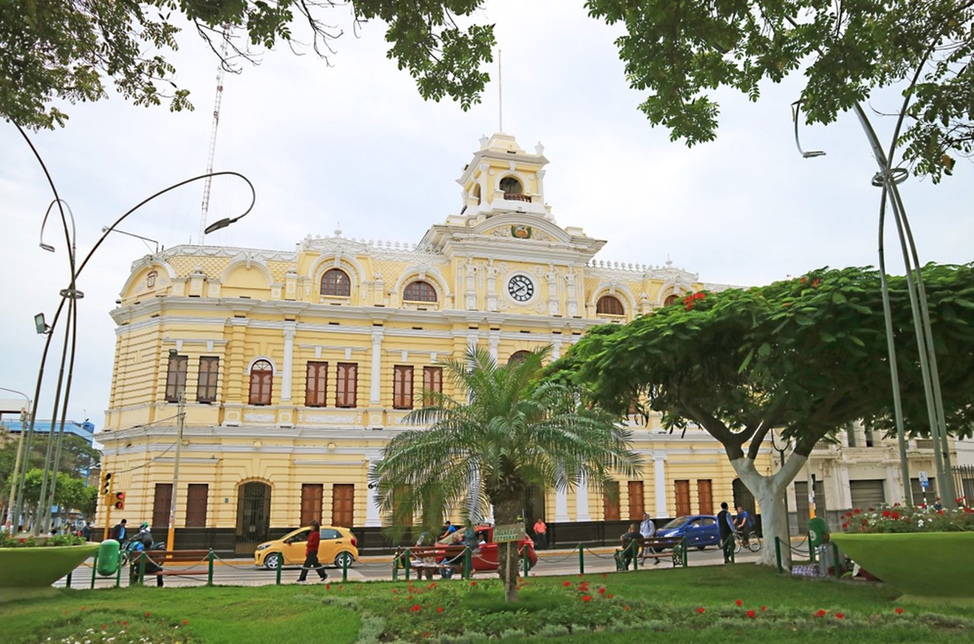 Geel historisch gebouw met klokgevel aan een plein in Peru, met bomen en passerend verkeer.