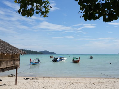 Strand in Thailand met longtailboten en speedboot in helderblauw water, met rieten hut op de voorgrond.