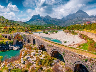 Oude stenen boogbrug over een turquoise rivier met bergen op de achtergrond in Albanië.