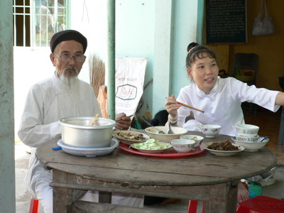 Traditionele Vietnamese maaltijd met twee mensen in witte kleding aan een tafel met schalen en eetstokjes.