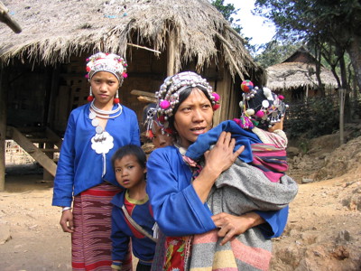 Vrouwen en kinderen in traditionele kleding bij een dorpshuis met rieten dak in Laos, Route Laos & Cambodja.